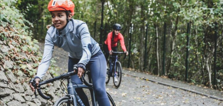 Two people are cycling on a cobblestone path through a forested area. They wear helmets, fitted athletic clothing, and are riding road bikes.