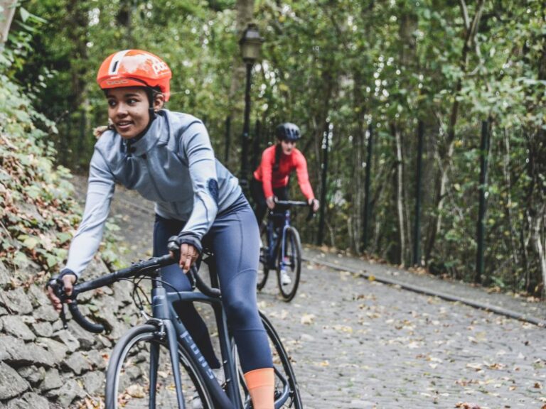 Two people are cycling on a cobblestone path through a forested area. They wear helmets, fitted athletic clothing, and are riding road bikes.