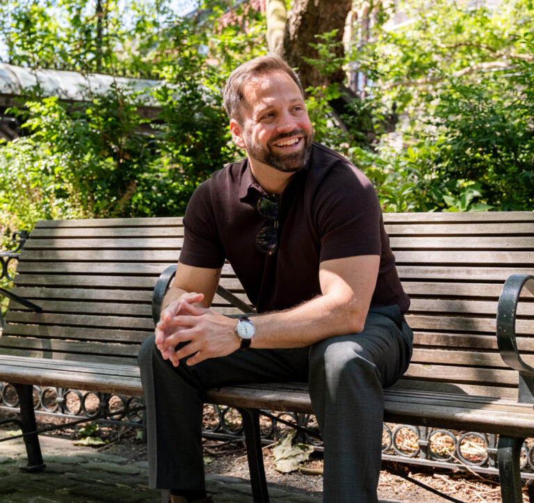 Adam smiles while sitting on a park bench, wearing a dark polo shirt, pants, watch, and sunglasses resting on the collar.