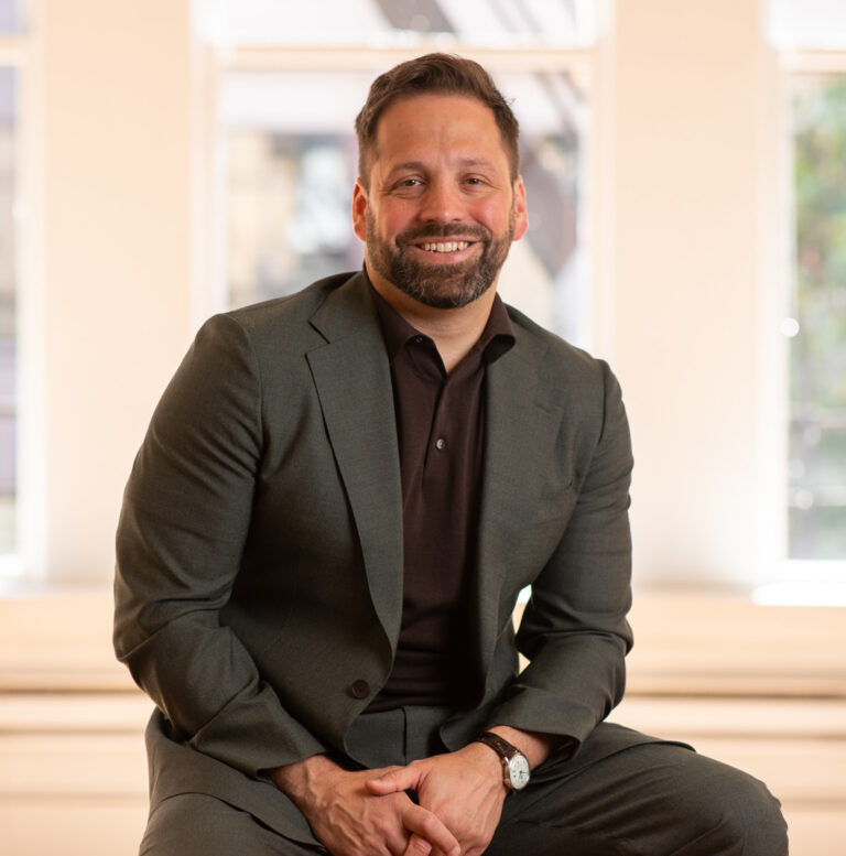 Adam smiles while seated, wearing a dark suit, and sporting a wristwatch in an indoor environment with natural light.