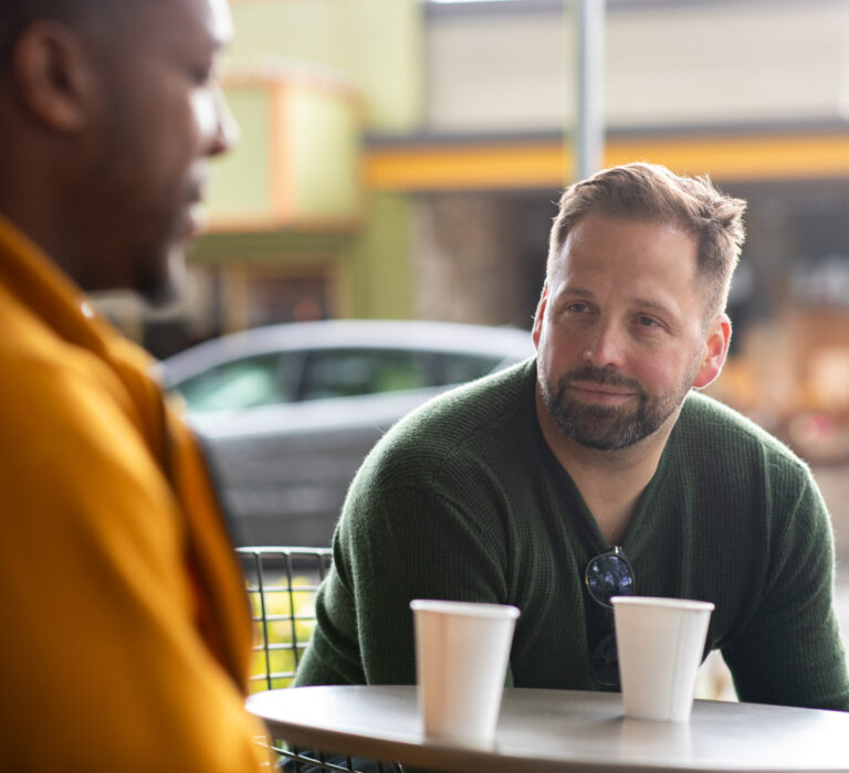 Adam and a Client are sitting across from each other at a table with coffee cups, having a conversation in a casual, daylight setting.