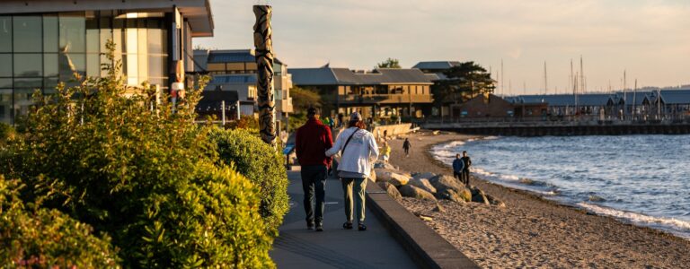 A seaside walkway in Edmond at dusk with people walking and enjoying the view, featuring a shoreline, greenery, and buildings in the background.