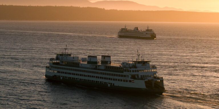 Two ferries cross in the Puget Sound at sunset, with the sun casting a warm glow over the mountains in the background.