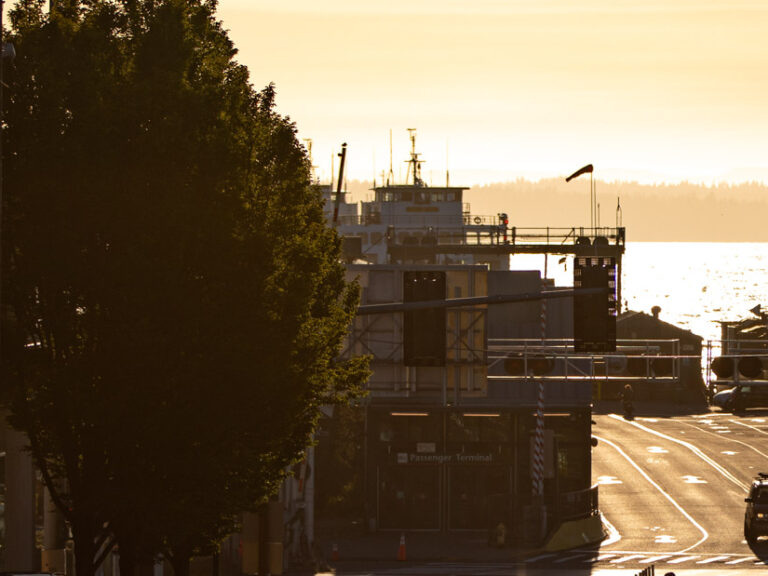 A serene sunset scene in Edmonds WA with street leading to a waterfront, with silhouettes of trees, vehicles, a ferry dock, and two people crossing the road.