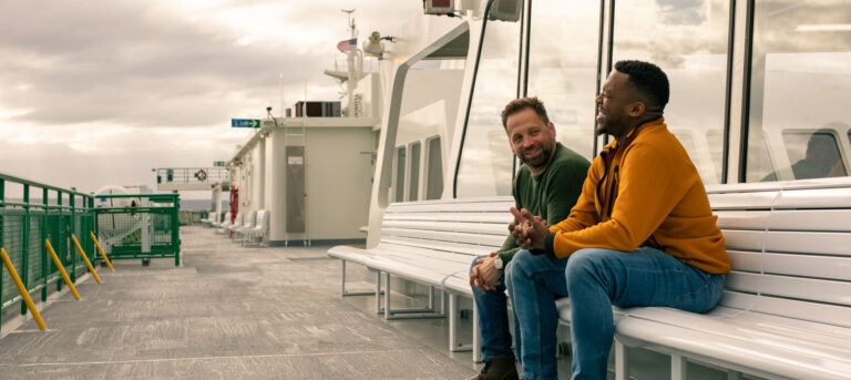 Adam and client are sitting on a ferry bench, talking and smiling. Overcast sky above, maritime setting, relaxed and cheerful atmosphere.