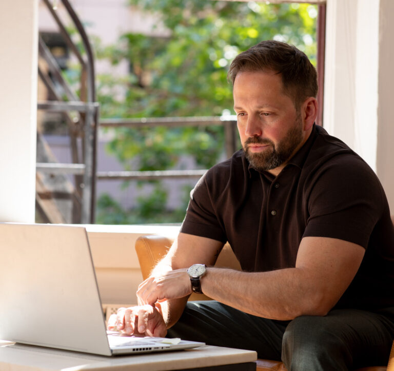 Adam at a table working on a laptop in a room with natural light and a view of greenery outside the window.