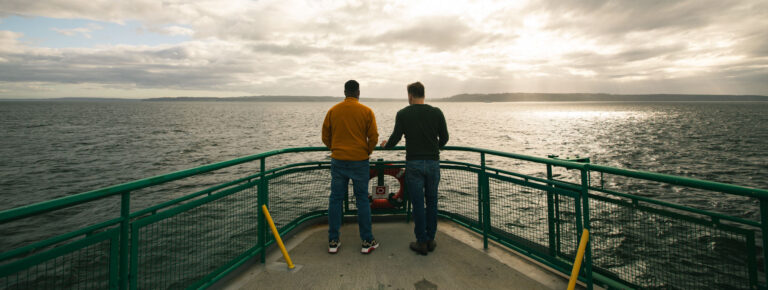 Adam and a client at the railing of a ferry, looking out at the Puget Sound under a cloudy sky. The horizon is visible in the distance.