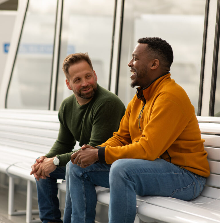 Adam and a client are sitting on a bench on a ferry, smiling and sharing a conversation. Adam is wearing a green sweater, the client a yellow zip up sweater.