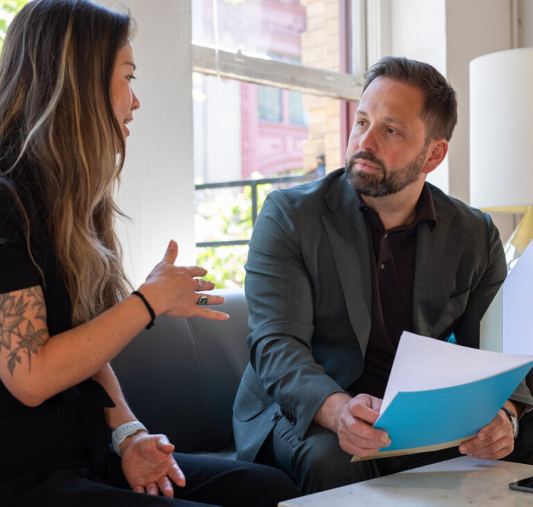 Adam and a client are in a conversation near a window. The client on the left gestures while speaking; Adam on the right listens, holding a folder.